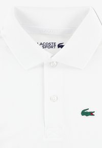 White polo shirt made of textured fabric, featuring a two-button placket and a green crocodile logo on the left chest.
