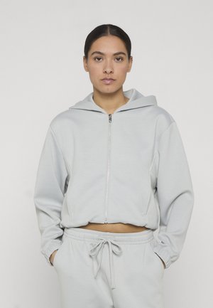 Sieviete stāv ar rokām kabatās, valkājot gaiši pelēku rāvējslēdzēja džemperi ar kapuci un atbilstošas bikses ar auklu, uz vienkārša fona.