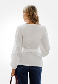 Blusa blanca ajustada con mangas largas tipo globo, cintura ceñida realzada por un cinturón blanco, combinada con jeans azul oscuro. Textura suave.