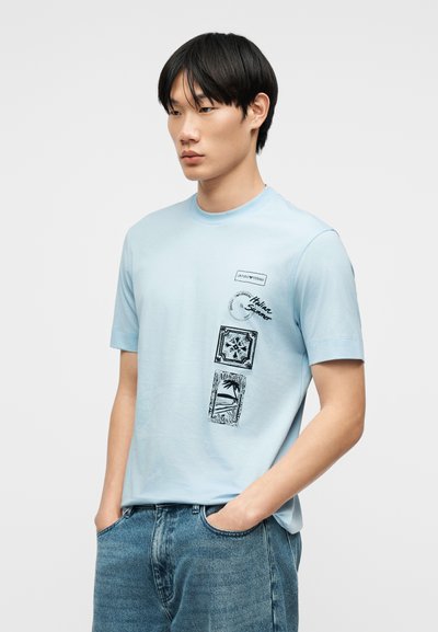 Νέος άνδρας που φοράει ανοιχτό μπλε μπλουζάκι με μαύρα γραφικά στη μέση και μπλε τζιν, στέκεται με τα χέρια στις τσέπες.