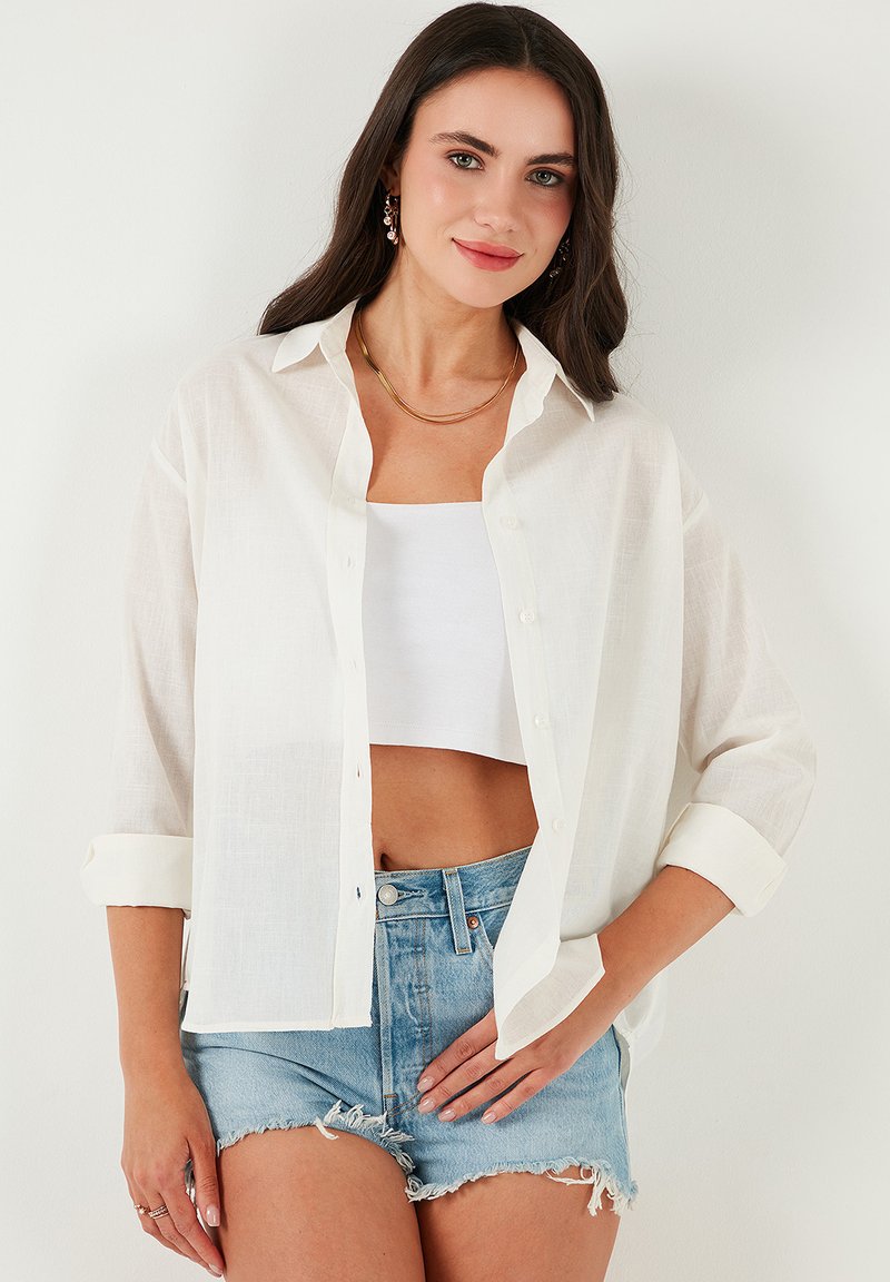 White linen shirt with a relaxed fit, button closure, and rolled sleeves, paired with a white crop top and distressed denim shorts.