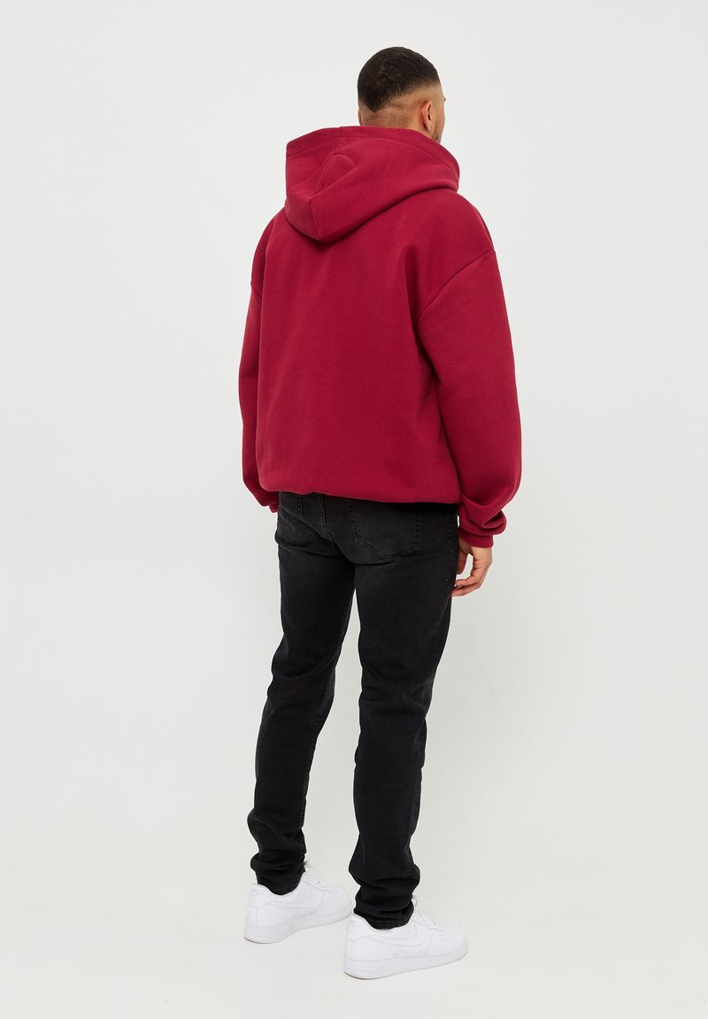 Hombre de pie de espaldas, vistiendo una sudadera con capucha roja, jeans negros y zapatillas blancas, contra un fondo claro y sencillo.