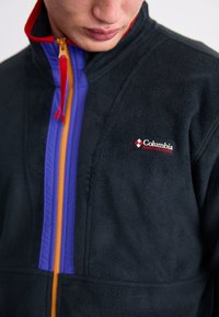 Čierna fleecová bunda s fialovým a oranžovým zipsom, s vysokým golierom a logom Columbia na hrudi. Mäkká textúra.