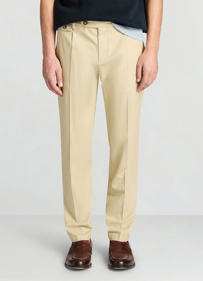 Pantalon beige à coupe ajustée, doté de doubles plis à l'avant et de deux boutons dorés à la taille, fabriqué dans un tissu lisse.
