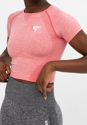 Lyhythihainen, istuva urheilupaita vaaleanpunaisesta kankaasta, jossa on kuvioitu tekstuuri, yhdistetty korkeavyötäröisiin tummanharmaisiin leggingsseihin, joissa on logo.