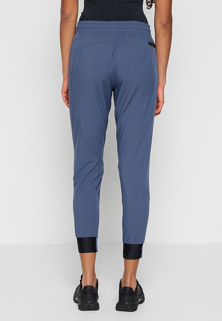 Femme portant un pantalon de sport bleu ajusté avec taille élastique et poignets noirs à la cheville, associé à des baskets noires, vue de derrière.