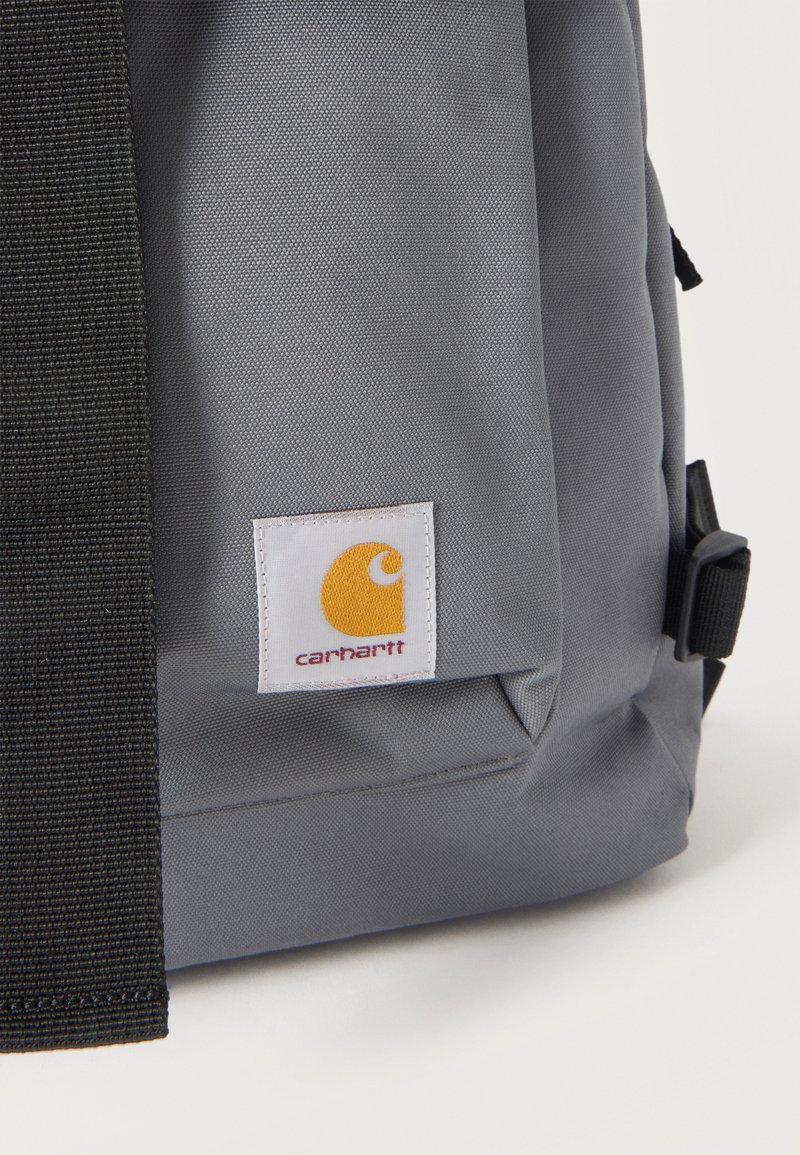Angolo dello zaino Carhartt grigio con una cinghia nera e un'etichetta bianca con il logo giallo Carhartt e testo rosso.