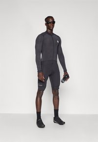 Bodysuit de sport noir à manches longues, design ajusté et fermeture éclair. Short de cyclisme court avec un logo. Lunettes de soleil noires et chaussures de cyclisme.