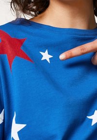 Camisa azul con una gran estrella roja y estrellas blancas de diferentes tamaños. El material parece suave con una textura lisa. Mano señalando el diseño.