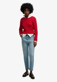 Kobieta z kręconymi włosami, ubrana w czerwoną sweterkową bluzę z splotem w warkocze, z białą koszulą pod spodem, jasnoniebieskie dżinsy i ciemnobrązowe mokasyny, stoi z rękami w kieszeniach.