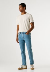 Beżowa koszulka z krótkim rękawem, jasnoniebieskie dżinsy i beżowe trampki. Koszulka ma okrągły dekolt i szczegóły logo na rękawie.