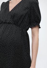 Schwarzes Kleid mit weißen Pünktchen, gepufften kurzen Ärmeln und V-Ausschnitt, getragen von einer Person vor einem einfarbigen hellen Hintergrund.