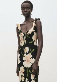 Zwart jurk met bloemenpatroon en ruches bij de schouders, versierd met lichtgekleurde bloemen en groene bladeren. Te combineren met een strik in de hals voor een verstelbare pasvorm.