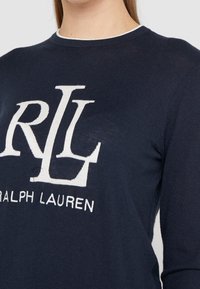 Kobieta w granatowym swetrze dziewiarskim z białym logo "RL" i napisem "Ralph Lauren" z przodu, skróconym na szyi i torsie.