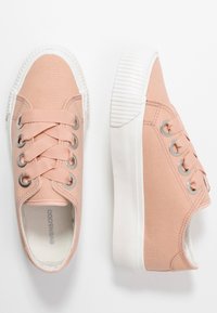 Jasnoróżowe płócienne trampki z białymi gumowymi podeszwami i szerokimi sznurówkami, przedstawione z widoku z góry i z boku na białym tle.