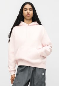 Femme aux longs cheveux foncés portant un sweat à capuche rose clair et un pantalon de jogging gris New Balance, debout avec une main dans la poche du sweat.