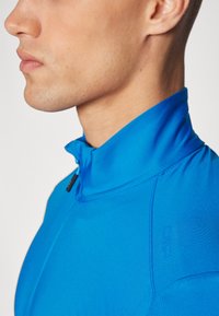 Blå idrettsjakke med glidelås og høy krage, laget av glatt stoff og med slim fit. Inkluderer en subtil logo på siden.