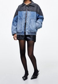 Chaqueta vaquera con diseño de bloques de color; paneles en azul claro y gris oscuro, mangas largas, botones a presión y bajo fruncido. Se lleva sobre una camiseta negra.