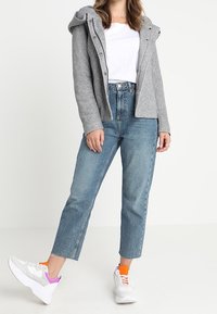 Grå huvjacka med knappar över en vit T-shirt, kombinerad med ljusblå jeans och vita sneakers med rosa och orange detaljer.