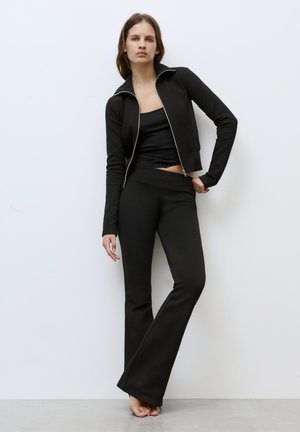 Žena oblékající černou bundu na zip, černé tílko a černé rozšířené kalhoty, stojící bosá proti bílé zdi.