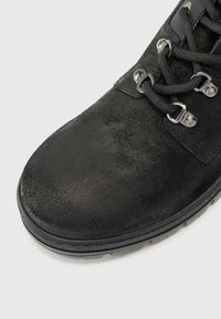 Prim-plan al unei pantofi din suede negru cu ochiuri din metal și șireturi negre, având o suprafață texturată și o talpă robustă.
