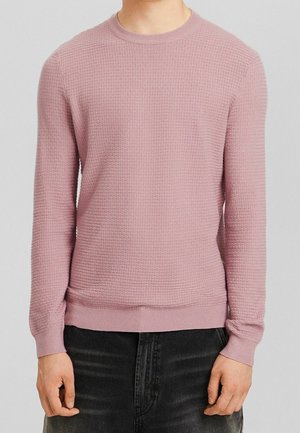 Mann trägt einen hellrosa strukturierten Langarmpullover kombiniert mit schwarzen Jeans, steht mit entspannten Armen seitlich.