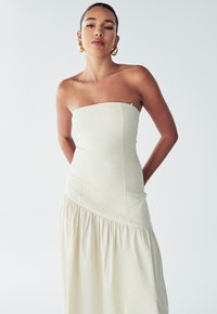 Robe crème sans bretelles avec un corsage ajusté et une jupe en couches fluide. Fabriquée en tissu léger, elle présente une texture subtile et des lignes épurées.
