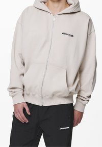 Sudadera con capucha beige con cremallera, bolsillo tipo canguro, puños y dobladillo acanalados. Presenta un pequeño logo negro en el área del pecho izquierdo. Textura de tela suave.