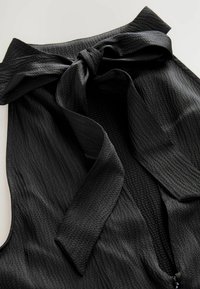 Schwarzes strukturiertes Kleidungsstück mit einer Schleifenverschluss. Es verfügt über eine weiche, gerippte Oberfläche und einen Reißverschluss, wobei der Fokus auf Einfachheit und Eleganz liegt.