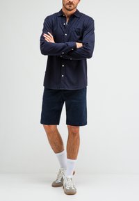 Marineblauwe button-up shirt met een zak, gecombineerd met donkere shorts. Het model draagt witte sokken en grijs-witte sneakers. Eenvoudige, casual uitstraling.