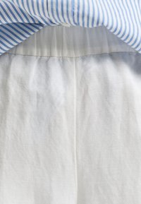 Col de chemise rayé bleu et blanc au-dessus du haut d’un pantalon blanc avec une couture visible à la ceinture.