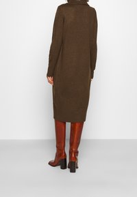 Robe tricotée marron avec un col montant, des manches longues et des fentes sur les côtés ; associée à des bottes marron brillantes à talons montant jusqu'aux genoux.