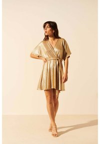 Robe dorée en métal avec des manches courtes, un col en V et une taille à nouer. Jupe évasée avec une texture brillante et longueur au-dessus du genou.