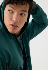 Sudadera verde con capucha y cuerda ajustable, con un corte relajado y un detalle gráfico negro en el frente, textura de algodón suave.