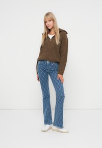 Brun strikket genser med hette, sammen med blå utsvingte jeans med diamantmønster og hvite sportssko med lys såle.