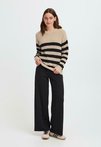 Maglione a righe beige e nero con vestibilità ampia, in tessuto a coste. Abbinato a pantaloni neri a gamba larga e sneakers beige. Design semplice.