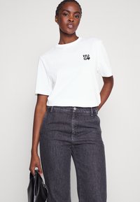 Valge lühikese varrukaga T-särk musta "HUGO" logoga, kombineeritud kõrge vöökohaga tumesiniste teksadega. Mudel hoiab musta kotti.