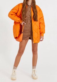 Veste matelassée orange en duvet avec deux poches, portée sur une robe à motifs marron. Complétée par des sneakers à plateforme crème et des chaussettes à motifs.