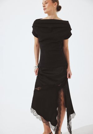 Donna che indossa un top nero scollato e una gonna nera asimmetrica con rifiniture in pizzo e spacco laterale, in piedi davanti a uno sfondo bianco.