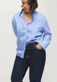 Camisa de botones azul claro con finas rayas verticales blancas, de mangas largas y corte relajado, combinada con jeans de mezclilla oscura.