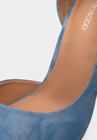 Κοντινό πλάνο μπλε υφασμάτινης ψηλοτάκουνης γόβας με ταμπά εσωτερική σόλα και λευκό λογότυπο της μάρκας στο εσωτερικό.