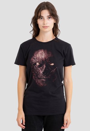 Young woman with wavy brown hair wearing a black T-shirt featuring a detailed, dark, monstrous face design.