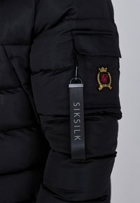 Sort pufferjakke med en lynlåslomme på ærmet, tekstureret stof og et mærke med "SIKSILK" i grå skrift.