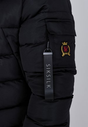 Giacca a vento nera con zip sulla manica, tessuto strutturato e un'etichetta con il marchio "SIKSILK" in lettere grigie.