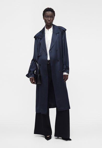 Άτομο που φορά σκούρο ναυτικό παλτό τύπου trench coat πάνω από λευκό πουκάμισο και μαύρο παντελόνι με φαρδιά μπατζάκια, κρατώντας μια μικρή μαύρη τσάντα με ουδέτερο φόντο.