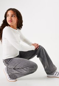 Witte geribde langesleevshirt met ronde hals, gecombineerd met loszittende grijze denim jeans en lichtgrijze sneakers met witte accenten.