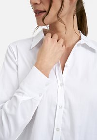White long-sleeve shirt with a button-down front, classic collar, and smooth fabric texture. Features clear buttons and wide cuffs.