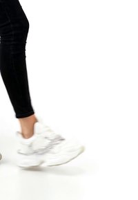 Persoană purtând pantaloni negri și adidași albi groși, mergând pe un fundal alb.