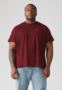 Bordeaux kortærmet t-shirt i et blødt, struktureret stof, med et lille hvidt logo på brystet, parret med lyseblå slidte jeans.