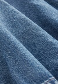 Denimstof med en medium blå nuance, der har en mærkbar tekstur og en let glans, og viser folder og rynker på overfladen.